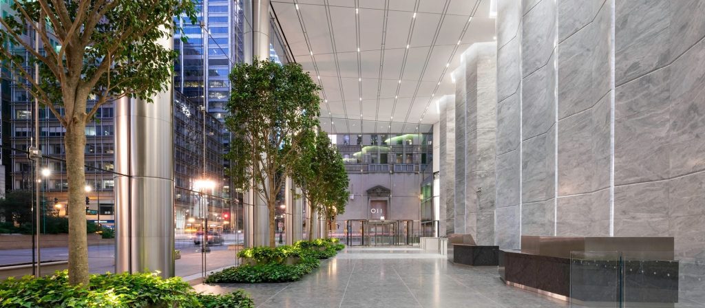 A spacious, modern lobby with tall marble walls, sleek metal columns, glass windows, and lush indoor greenery in the center of Chicago - YOU Psychiatry Clinic
